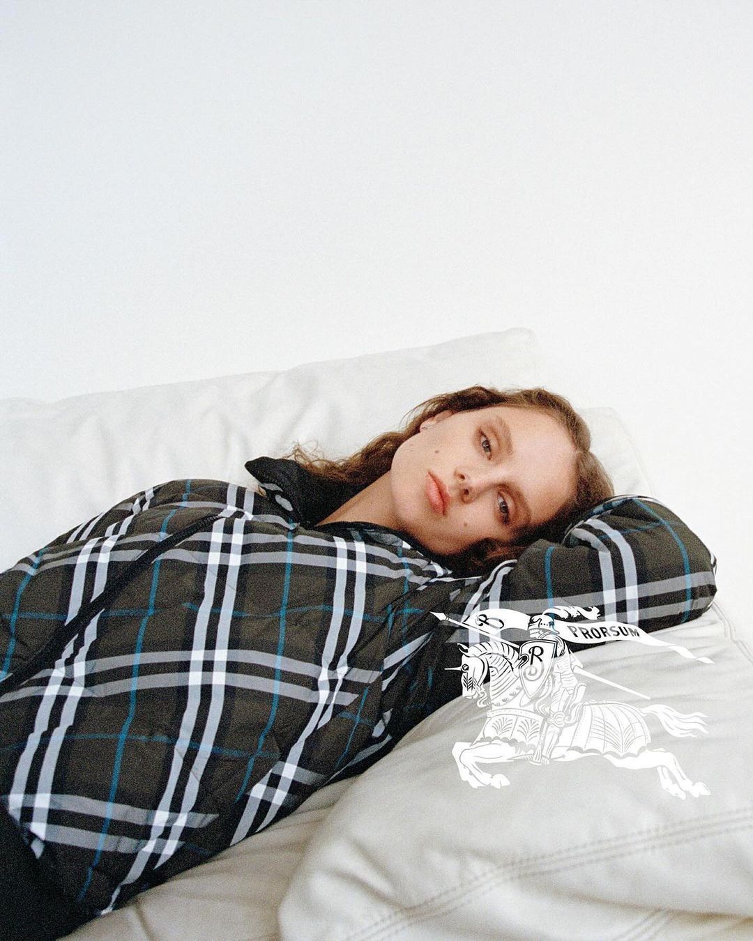 A young woman lies on a white cushion wearing a plaid jacket with a brand logo, against a white background in a studio environment.