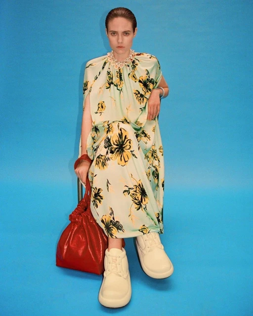 A woman in a floral dress poses against a blue backdrop, holding a red bag and wearing platform shoes in a fashion-forward editorial shot.