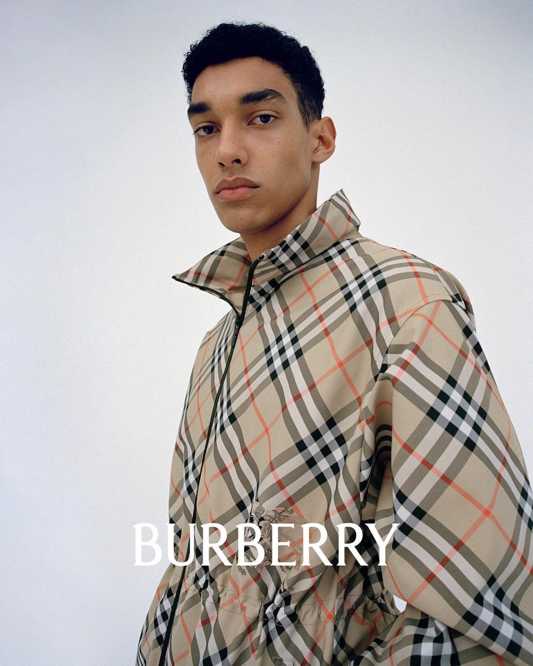 Portrait of a slender adult male wearing a Burberry plaid zip jacket against a white background, showcasing high-fashion apparel.