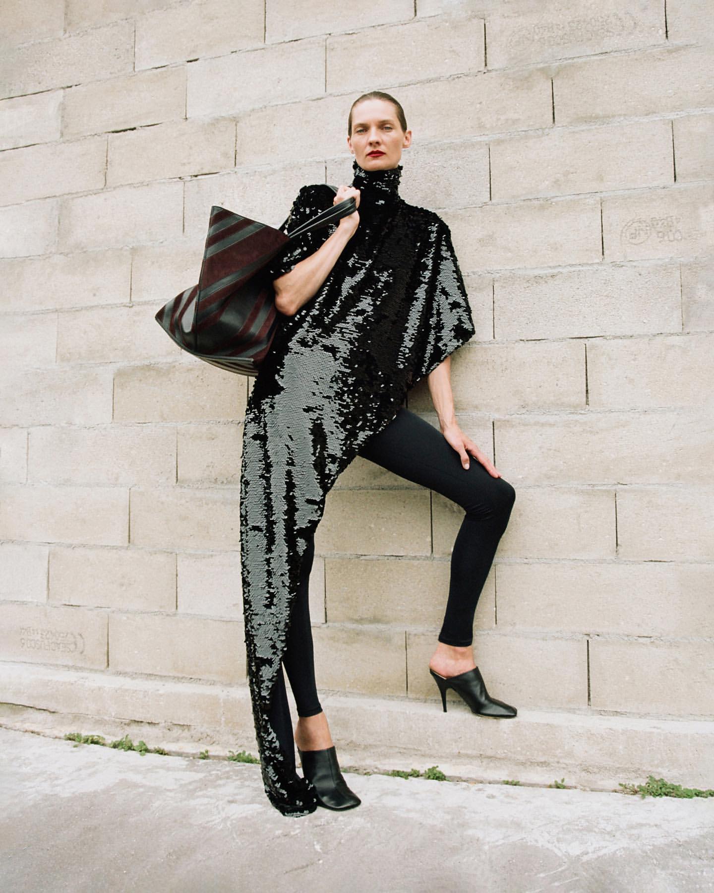 Fashion model in a sequined asymmetrical top, black leggings, and heels poses against a stone brick wall with a large tote bag.