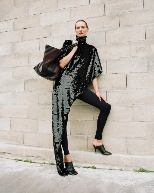 Fashion model in a sequined asymmetrical top, black leggings, and heels poses against a stone brick wall with a large tote bag.
