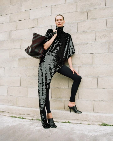 Fashion model in a sequined asymmetrical top, black leggings, and heels poses against a stone brick wall with a large tote bag.