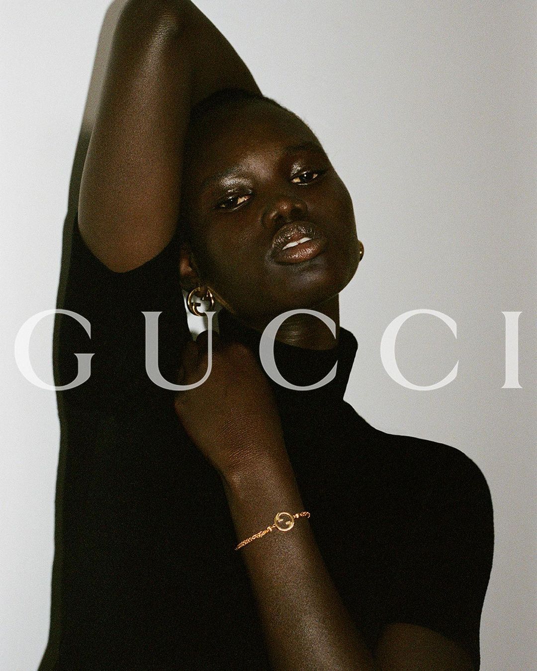 A dark-skinned woman poses in a close-up fashion shot with gold jewelry, a black turtleneck, and a background featuring the GUCCI logo.