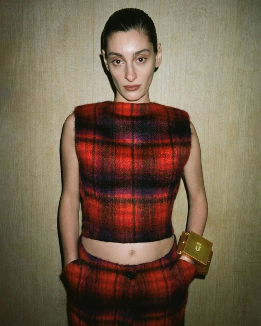 A stylish young woman poses indoors wearing a red and black plaid sleeveless top and matching pants, accessorized with a gold bracelet.