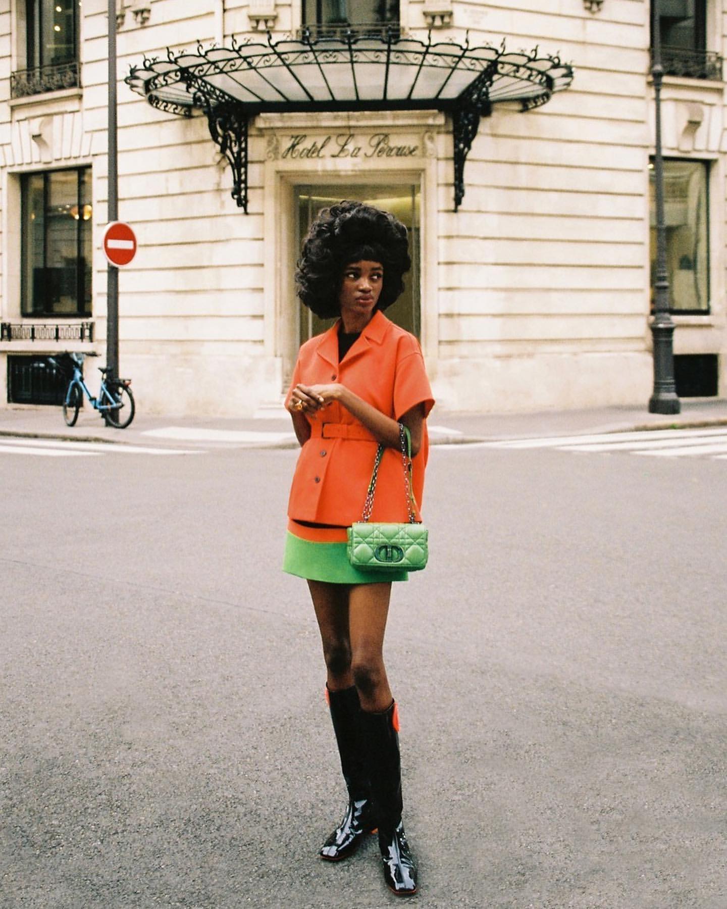 A stylish woman poses in an orange jacket and green skirt on a city street near a hotel, creating a fashion-forward, urban portrait.