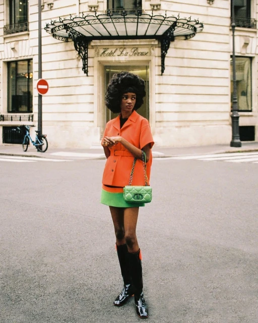A stylish woman poses in an orange jacket and green skirt on a city street near a hotel, creating a fashion-forward, urban portrait.