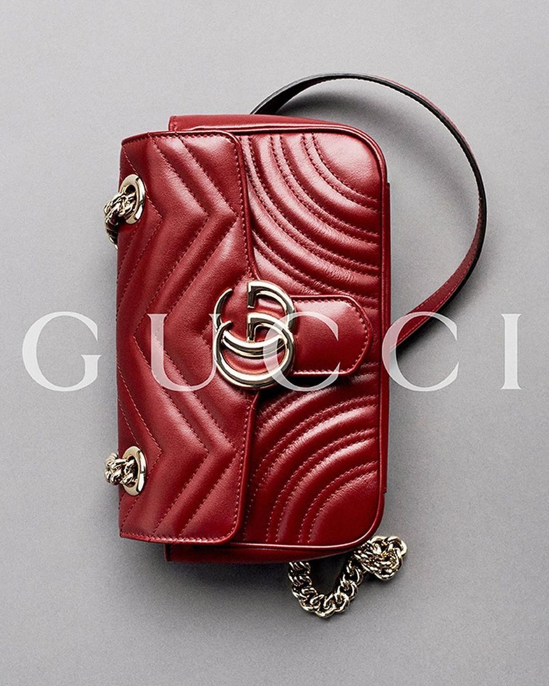 Close-up studio shot of a red leather Gucci Marmont handbag with chevron quilting, chain strap, and signature metal logo on a gray background.