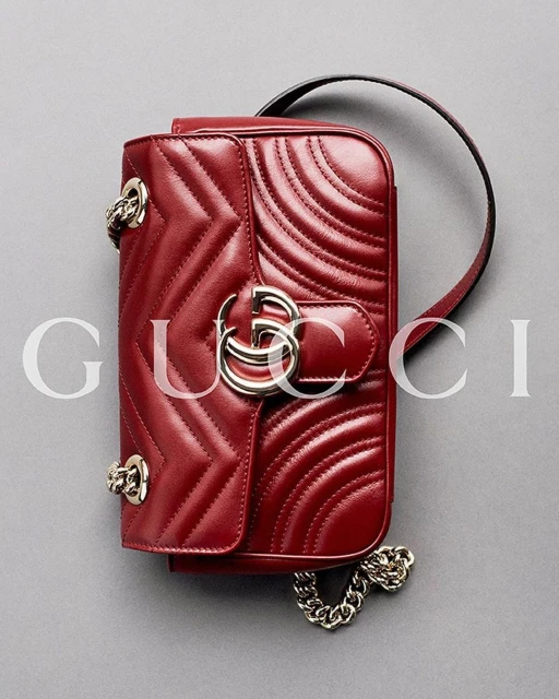 Close-up studio shot of a red leather Gucci Marmont handbag with chevron quilting, chain strap, and signature metal logo on a gray background.
