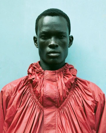 Portrait of a Black man with a serious expression wearing a red gathered jacket against a light blue wall.