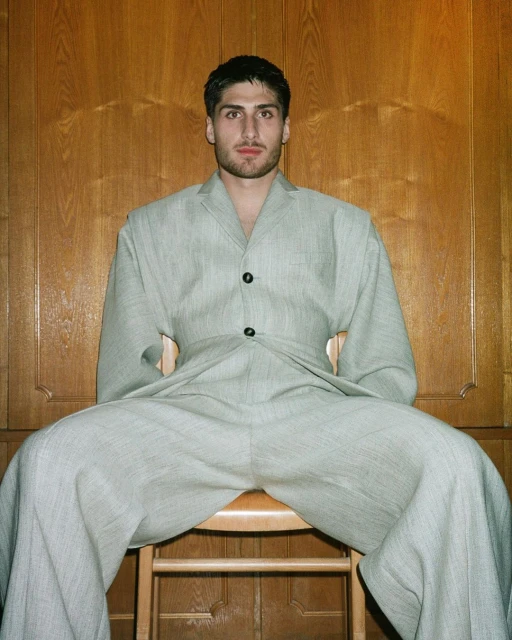Man with fair skin sits on a wooden chair in a light green-gray suit, wooden wall panel background. Indoor fashion shot.