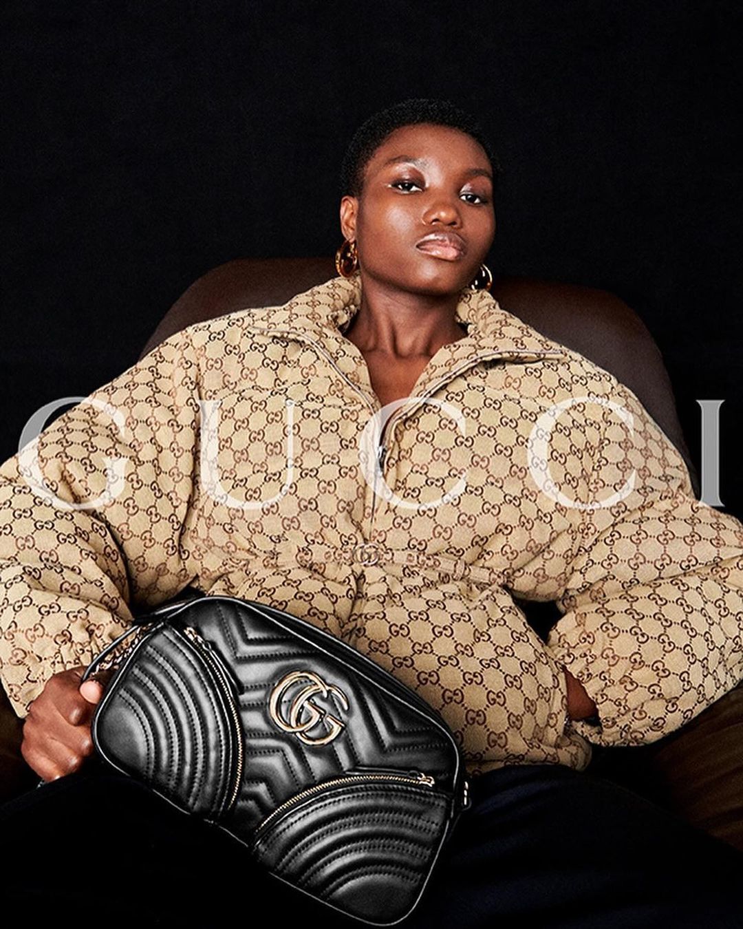 A young woman models in a studio wearing a beige Gucci-print jacket, seated with a black Gucci handbag on her lap.