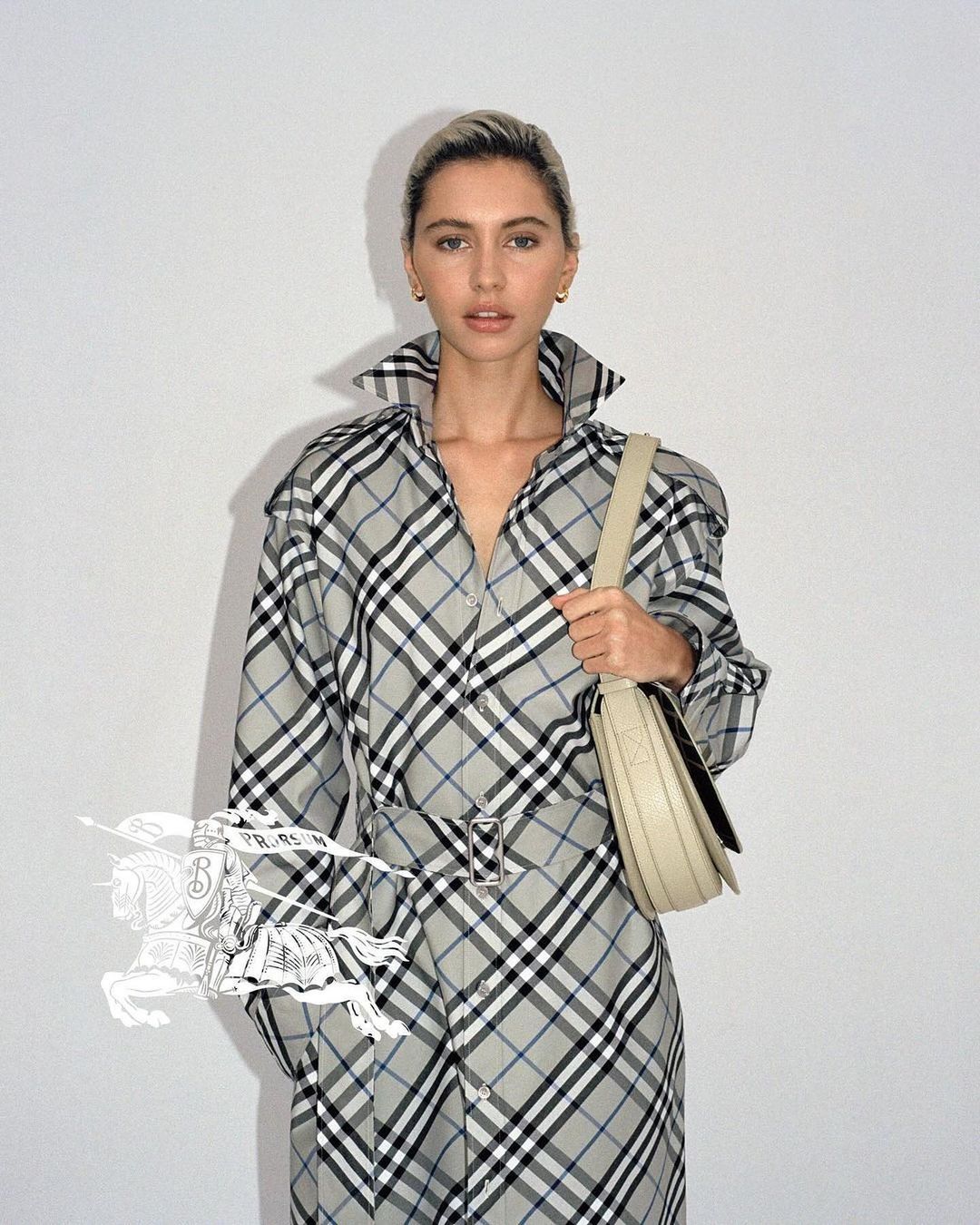 Woman in Burberry plaid trench coat poses indoors against a white wall, holding a light-beige leather shoulder bag.