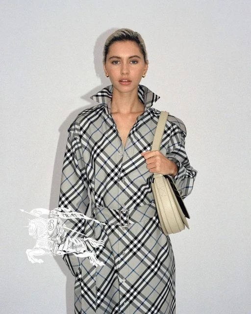Woman in Burberry plaid trench coat poses indoors against a white wall, holding a light-beige leather shoulder bag.