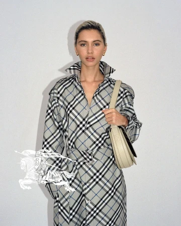 Woman in Burberry plaid trench coat poses indoors against a white wall, holding a light-beige leather shoulder bag.
