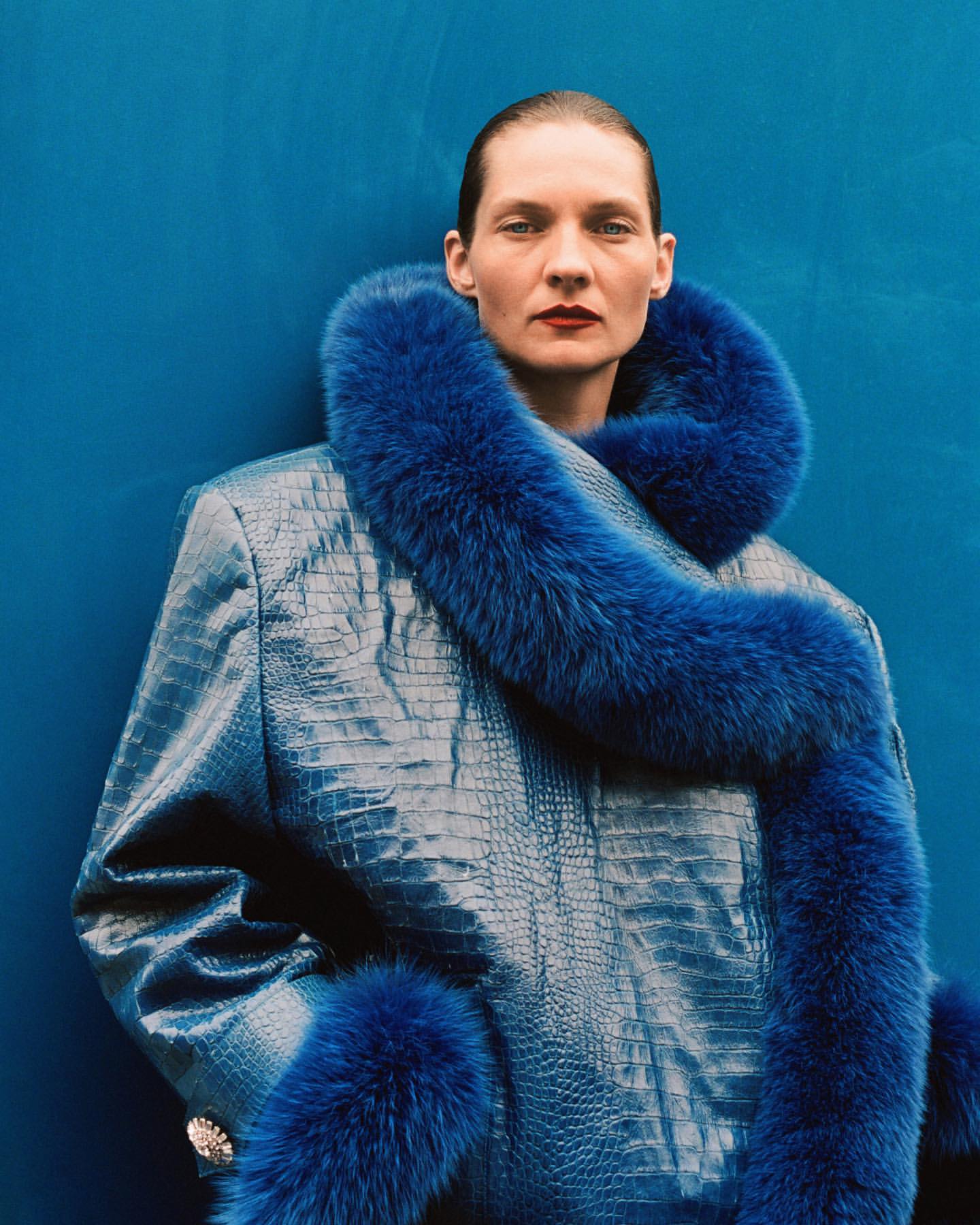 Elegant adult woman in a blue jacket with fur trim against a solid blue background in a studio fashion shot. Shoulder length, portrait.
