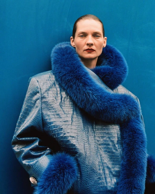 Elegant adult woman in a blue jacket with fur trim against a solid blue background in a studio fashion shot. Shoulder length, portrait.