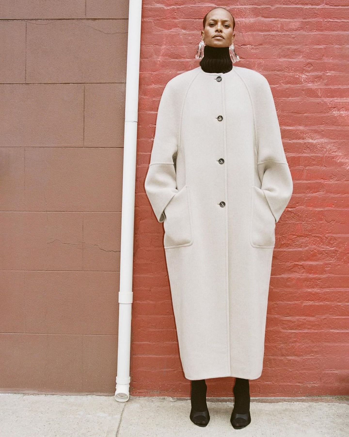 Elegant woman in a cream-colored coat poses against a red brick wall wearing black turtleneck, earrings, and black heels in a studio shot.