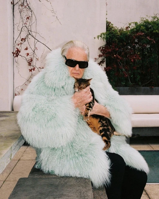 Elderly adult and tabby cat, posing outdoors in a plush mint-green coat and dark sunglasses, on stone steps, sunny daytime.