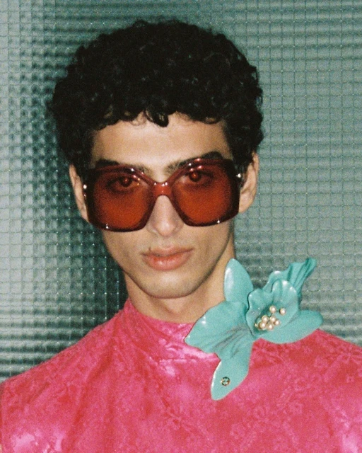 A person with dark, curly hair wearing red glasses and a pink shirt poses indoors against a textured background with a floral brooch.