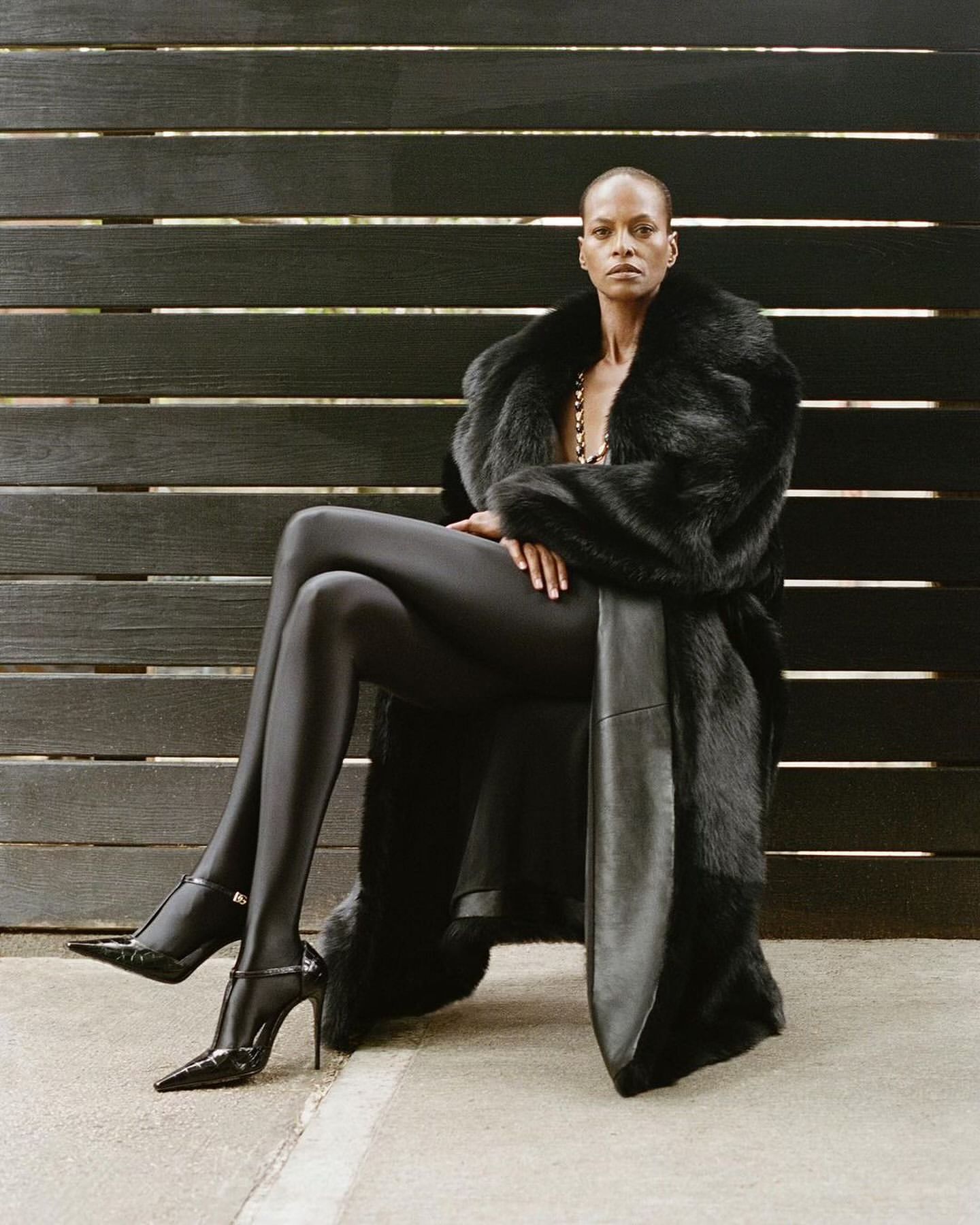 Elegant, bald, Black woman sits in a faux fur coat, glossy leggings and black heels. She's posing against a dark wood plank wall.