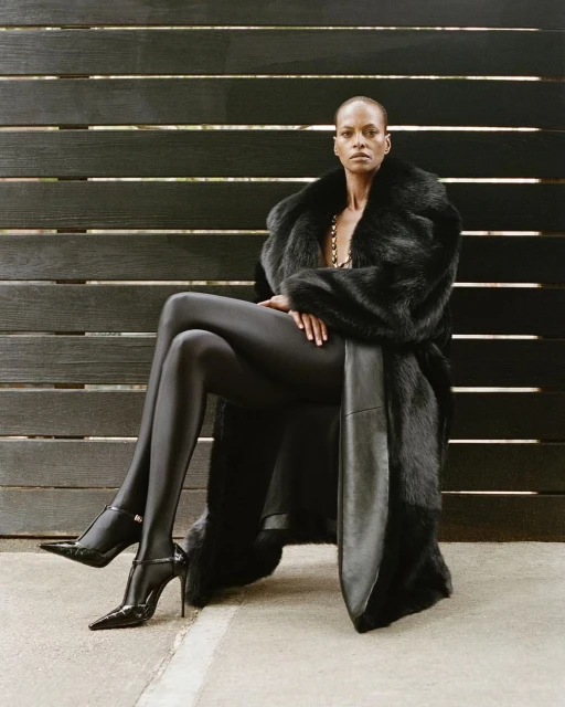 Elegant, bald, Black woman sits in a faux fur coat, glossy leggings and black heels. She's posing against a dark wood plank wall.