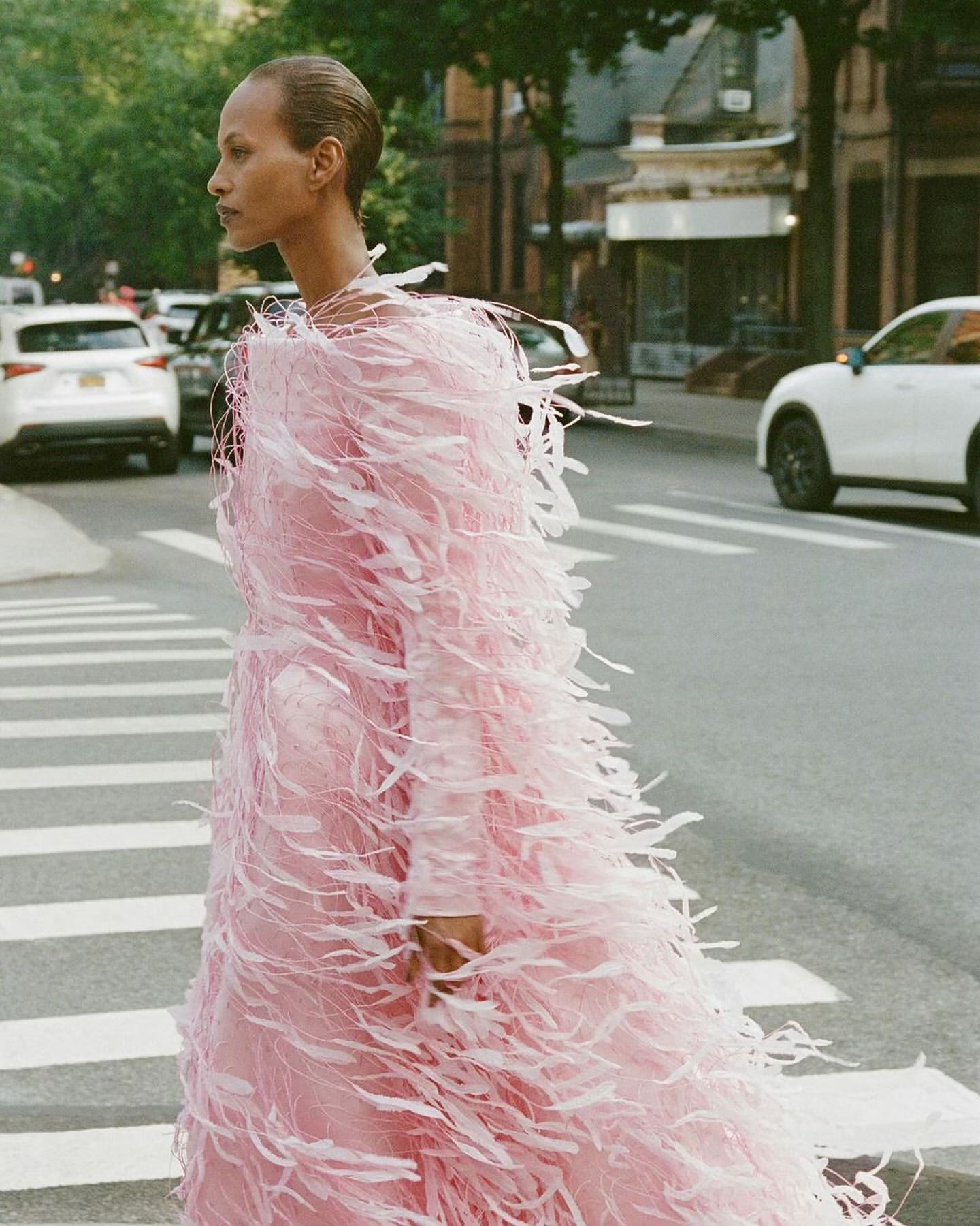 Elegant woman in a pink feathered dress walks confidently on a zebra crossing in a city street, surrounded by vehicles and buildings.