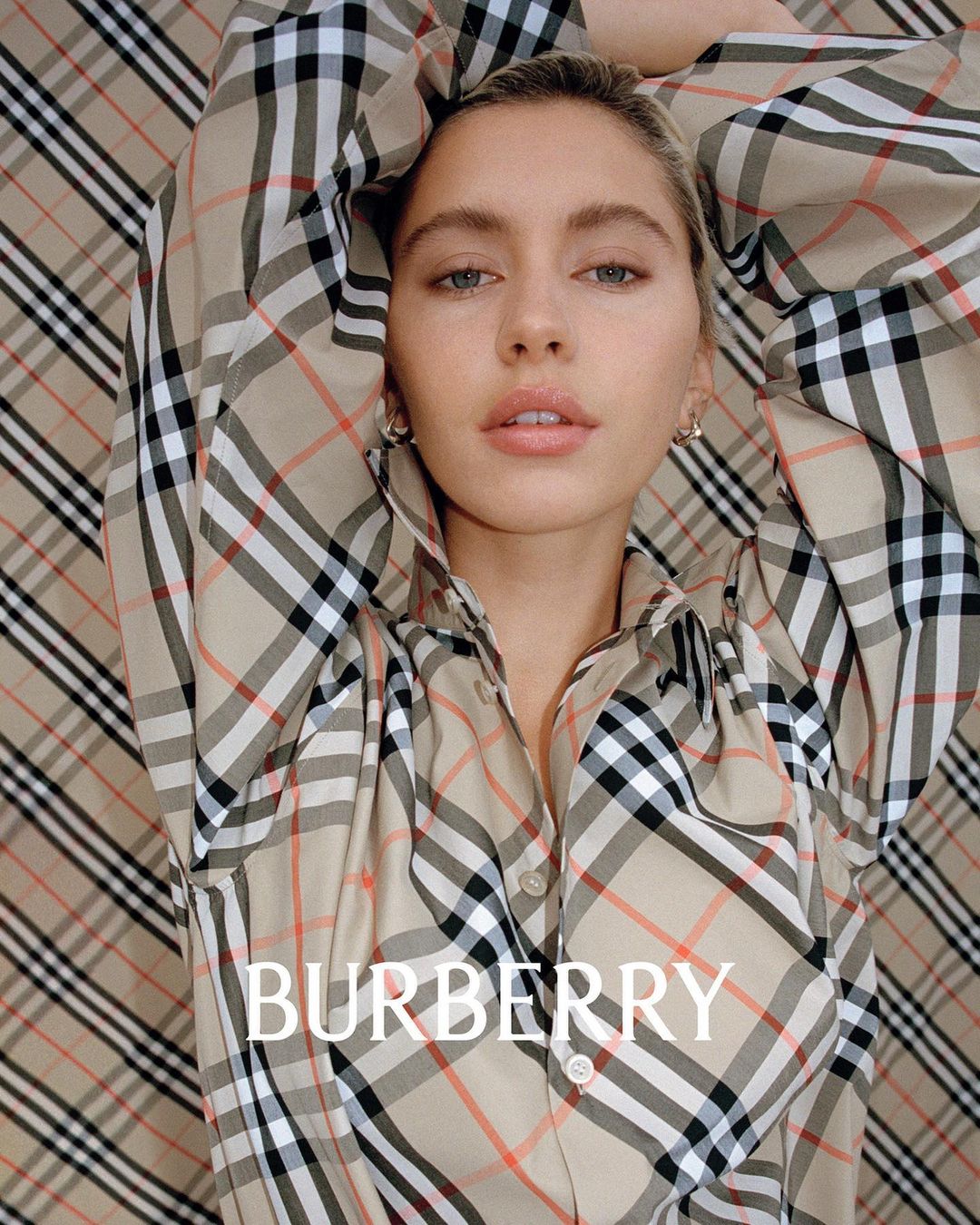 Blonde woman poses against a Burberry pattern backdrop, wearing a Burberry plaid button-down shirt, styled for a high-fashion editorial shot.