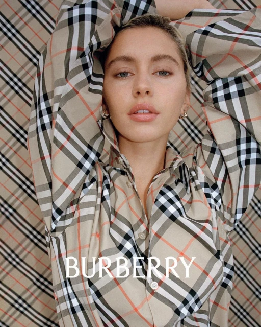 Blonde woman poses against a Burberry pattern backdrop, wearing a Burberry plaid button-down shirt, styled for a high-fashion editorial shot.