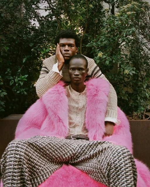 Two dark-skinned models pose outdoors: one seated in a pink fur coat with patterned pants, and one standing behind wearing a knit sweater.