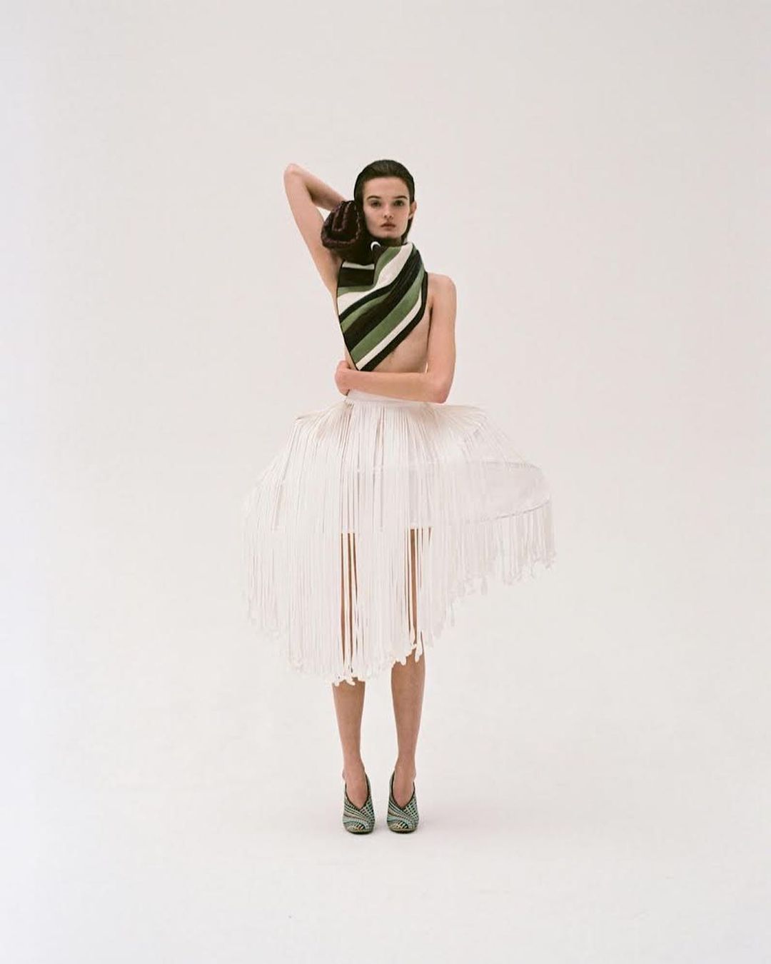 Model wearing a striped scarf and structured white fringed skirt standing against a white seamless background