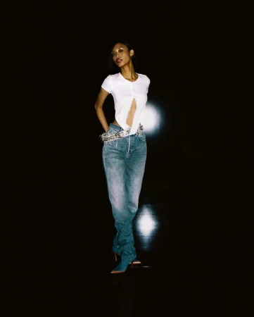 Model standing against a dark background wearing a white top and denim jeans by Davit Giorgadze