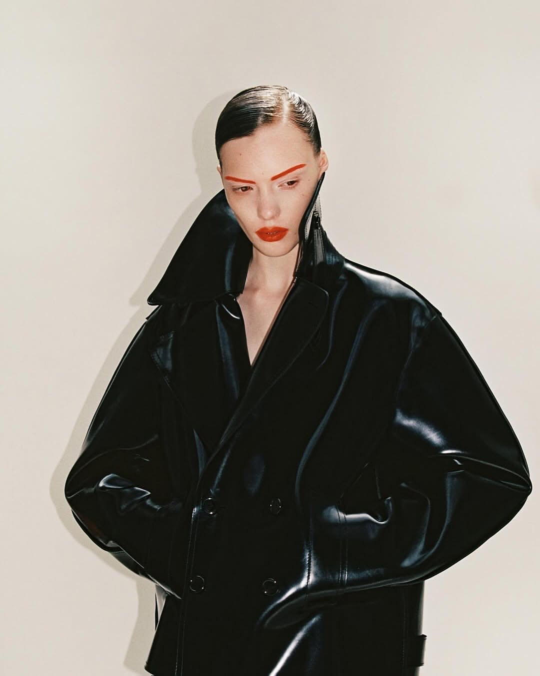 Close-up studio shot of a female fashion model wearing an oversized black vinyl trench coat with dramatic red makeup against a white backdrop.