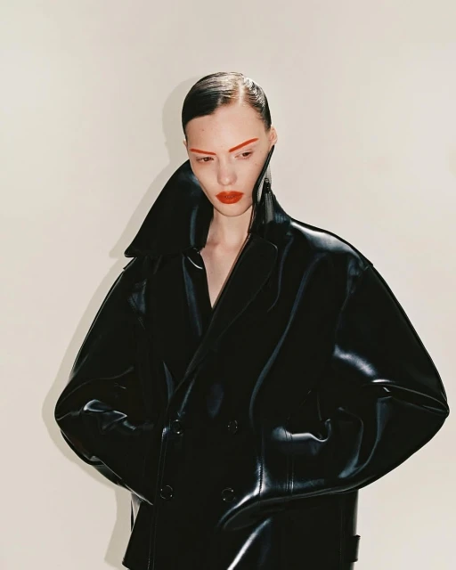 Close-up studio shot of a female fashion model wearing an oversized black vinyl trench coat with dramatic red makeup against a white backdrop.