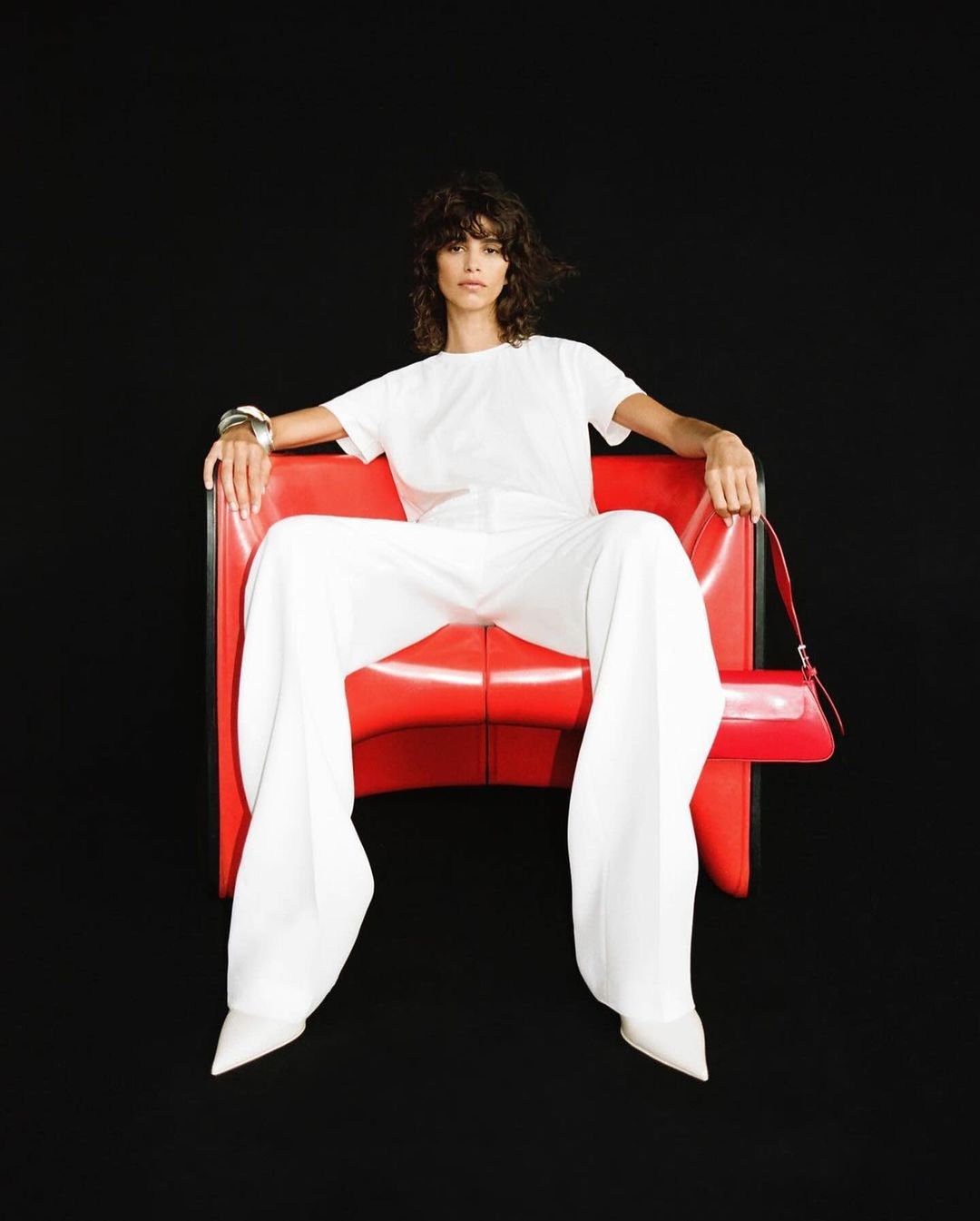 A woman with curly hair is sitting in a red chair wearing a white t-shirt and wide-legged pants, posing against a dark background.