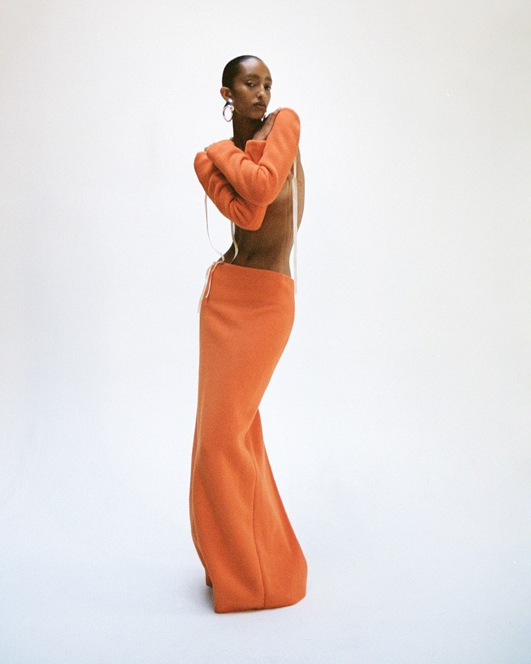 A Black female model in an orange knit skirt and arm warmers poses in a full shot against a white backdrop in a fashion setting.