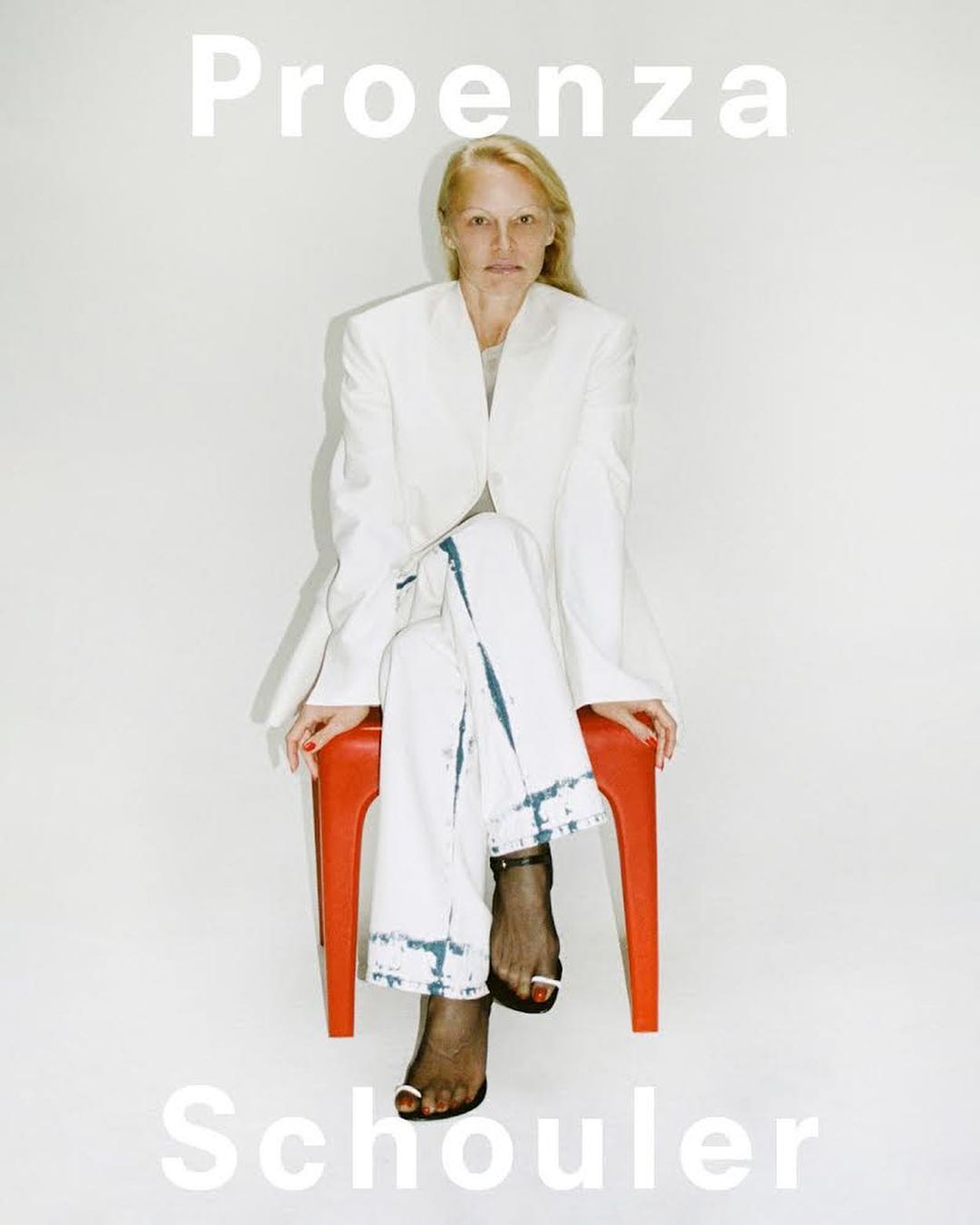 A blonde woman in a white blazer and pants sits on an orange stool in a studio setting for a Proenza Schouler fashion campaign.