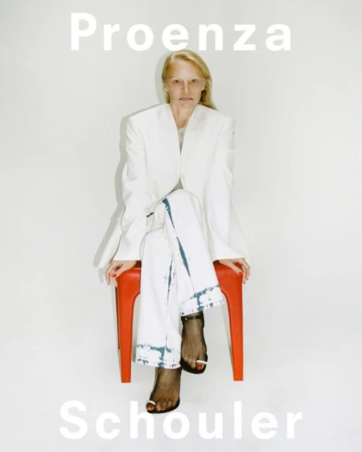 A blonde woman in a white blazer and pants sits on an orange stool in a studio setting for a Proenza Schouler fashion campaign.