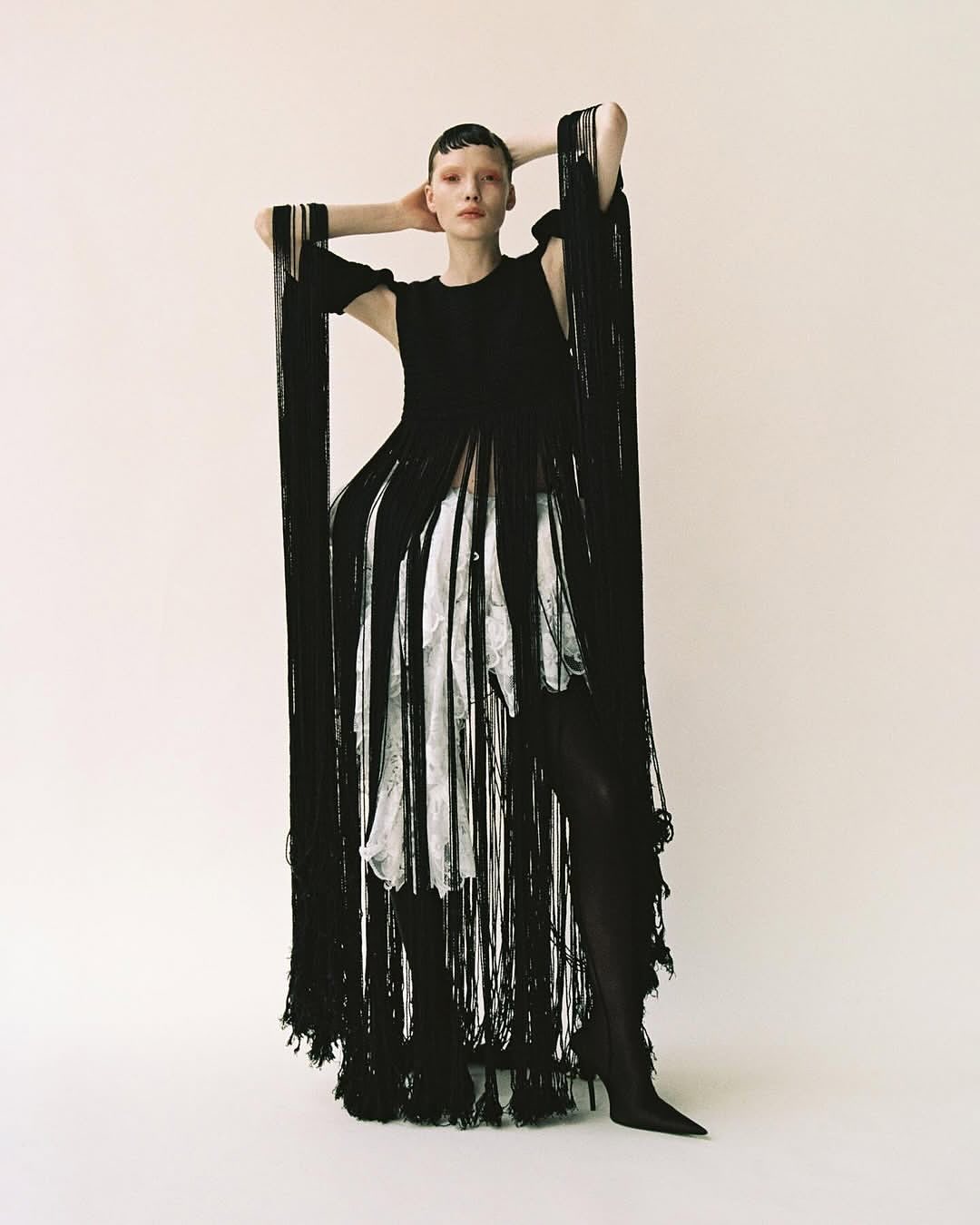 Model with short dark hair in black fringed dress and white patterned underlayer standing against a neutral studio backdrop