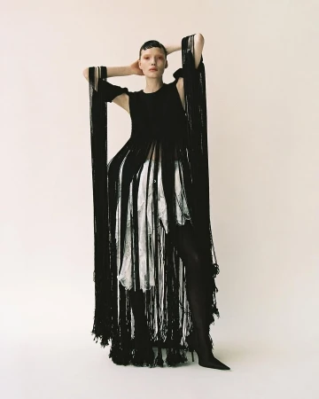 Model with short dark hair in black fringed dress and white patterned underlayer standing against a neutral studio backdrop