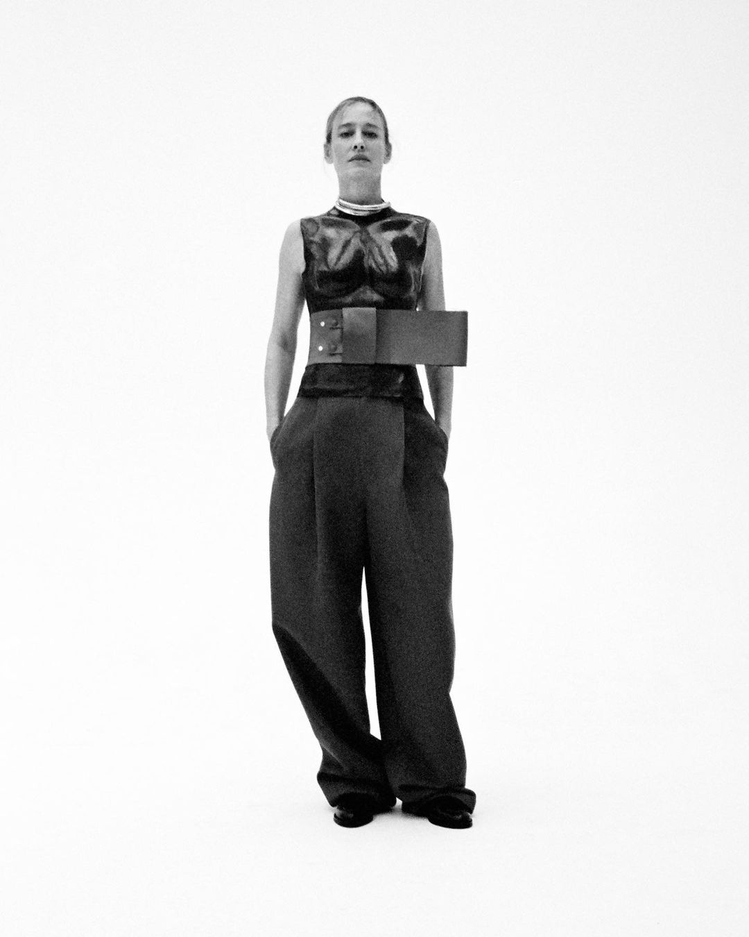 Monochrome fashion portrait of a model wearing a sleeveless dark top and wide trousers against a white background.