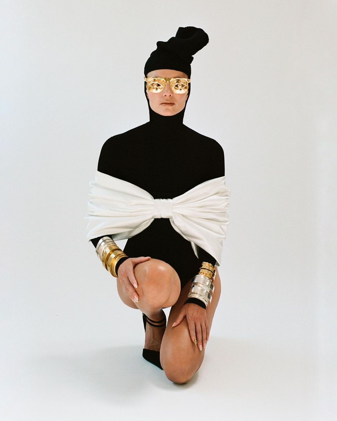 A female fashion model in gold sunglasses, black bodysuit, and silver and gold bracelets poses kneeling against a plain white backdrop.