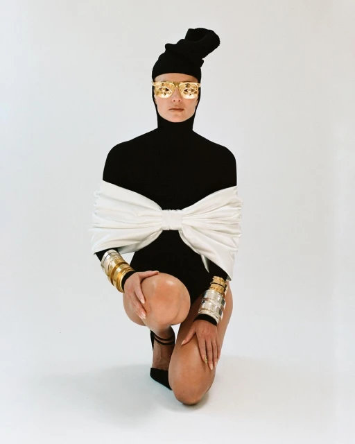 A female fashion model in gold sunglasses, black bodysuit, and silver and gold bracelets poses kneeling against a plain white backdrop.