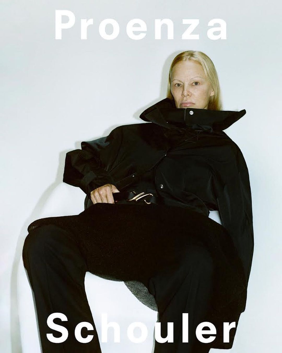 Model wearing a black jacket and dark trousers seated against a plain white background with bold white typography.