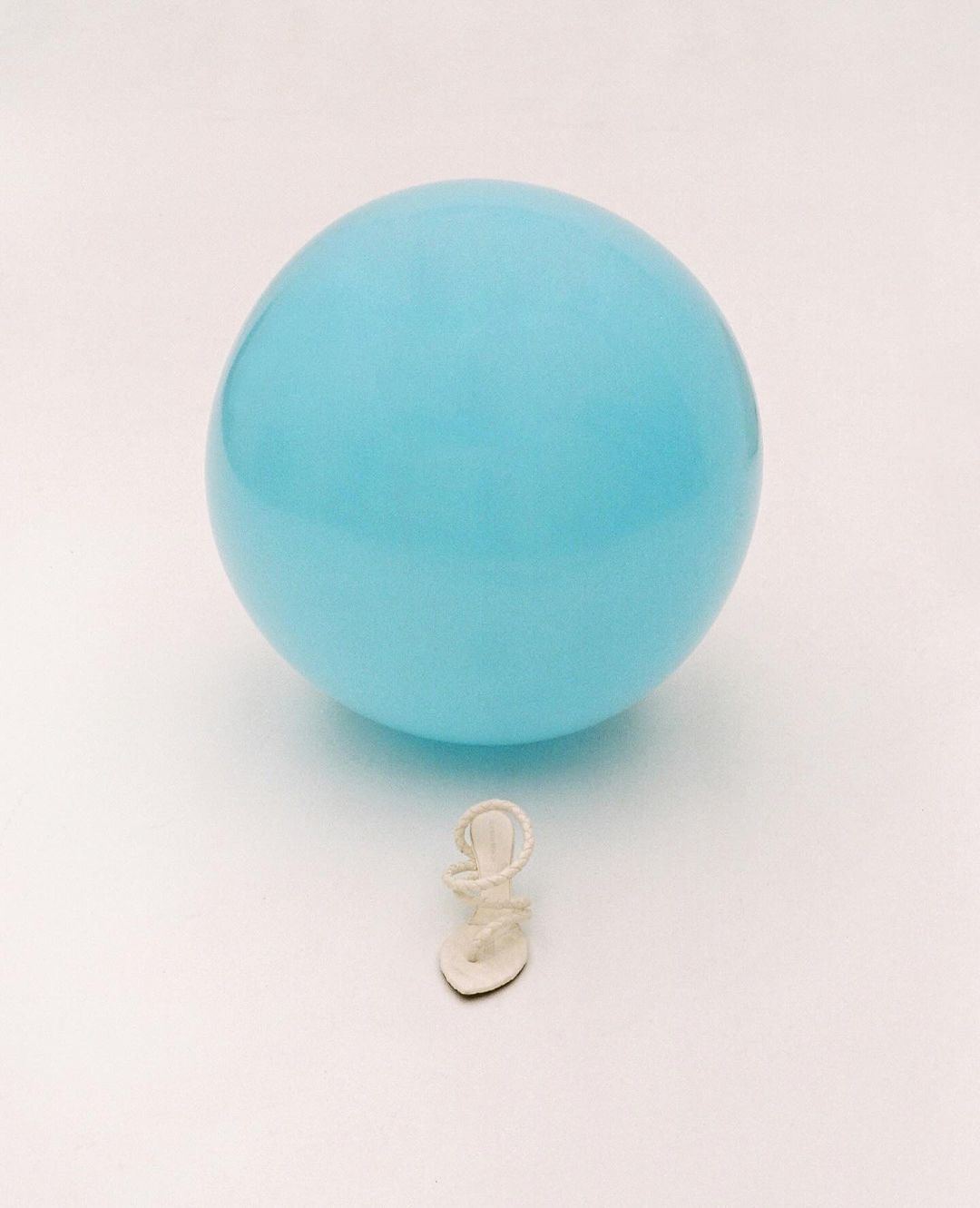 Close-up shot of a single white strappy sandal placed beneath a light-blue balloon, set against a clean, bright white backdrop.