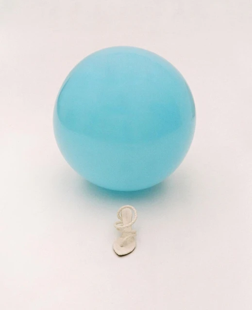 Close-up shot of a single white strappy sandal placed beneath a light-blue balloon, set against a clean, bright white backdrop.