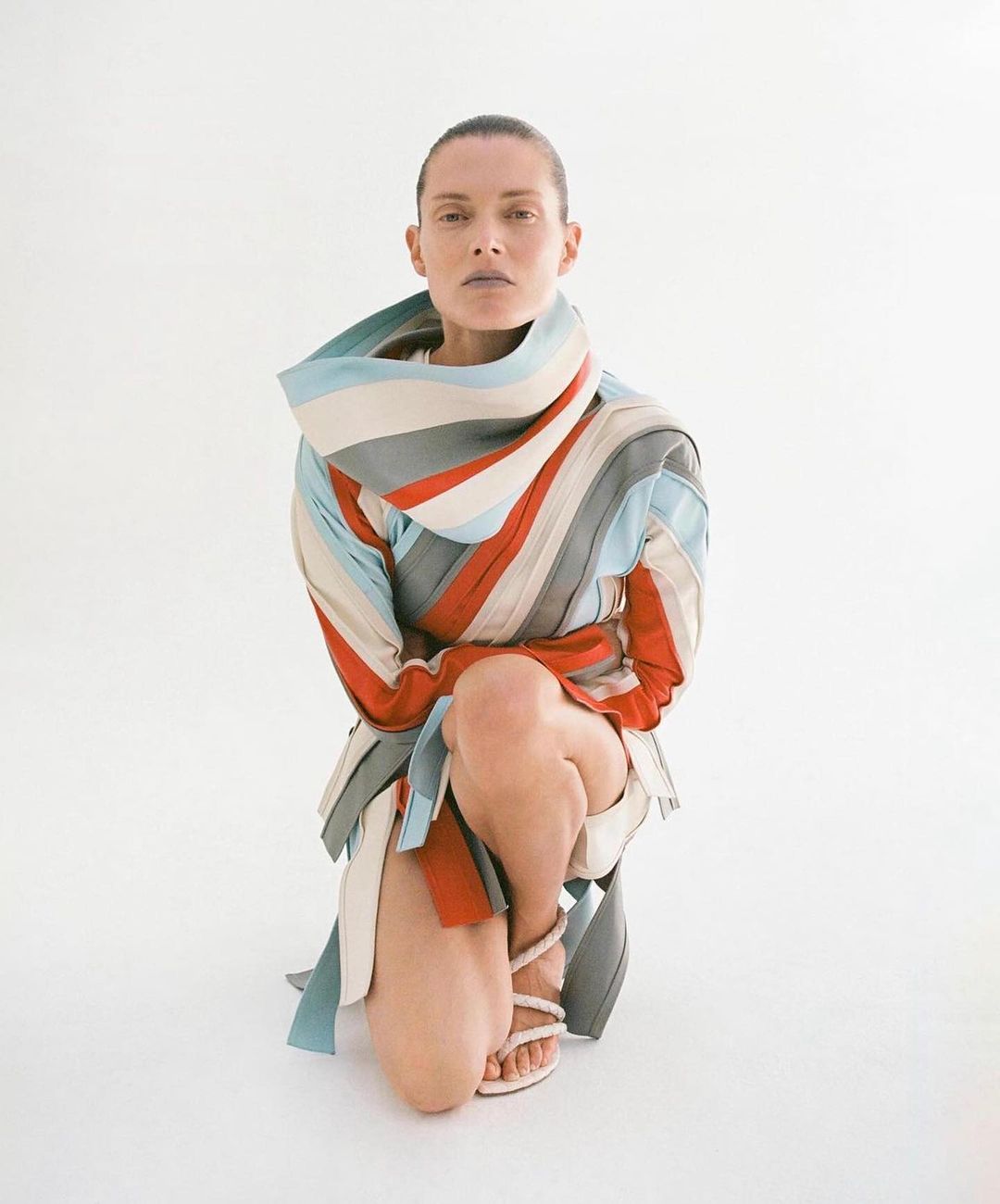 Woman kneeling in a studio setting wearing a striped garment and strappy heels. She has a neutral expression and clean shaven head.