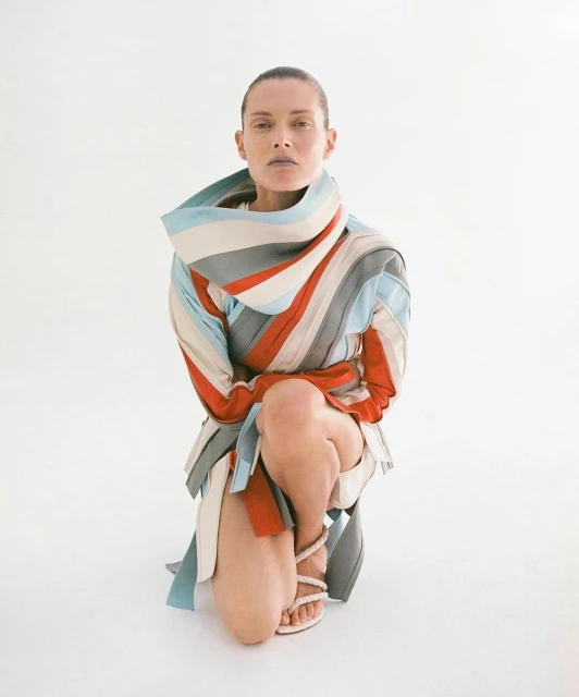 Woman kneeling in a studio setting wearing a striped garment and strappy heels. She has a neutral expression and clean shaven head.