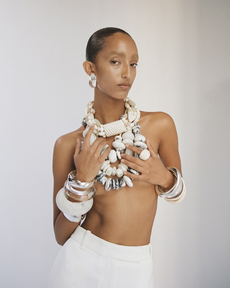 Woman poses in a studio with light skin and short hair wearing a white top and large handmade jewelry including a necklace and bracelets.
