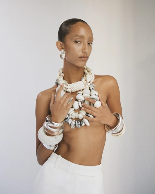 Woman poses in a studio with light skin and short hair wearing a white top and large handmade jewelry including a necklace and bracelets.