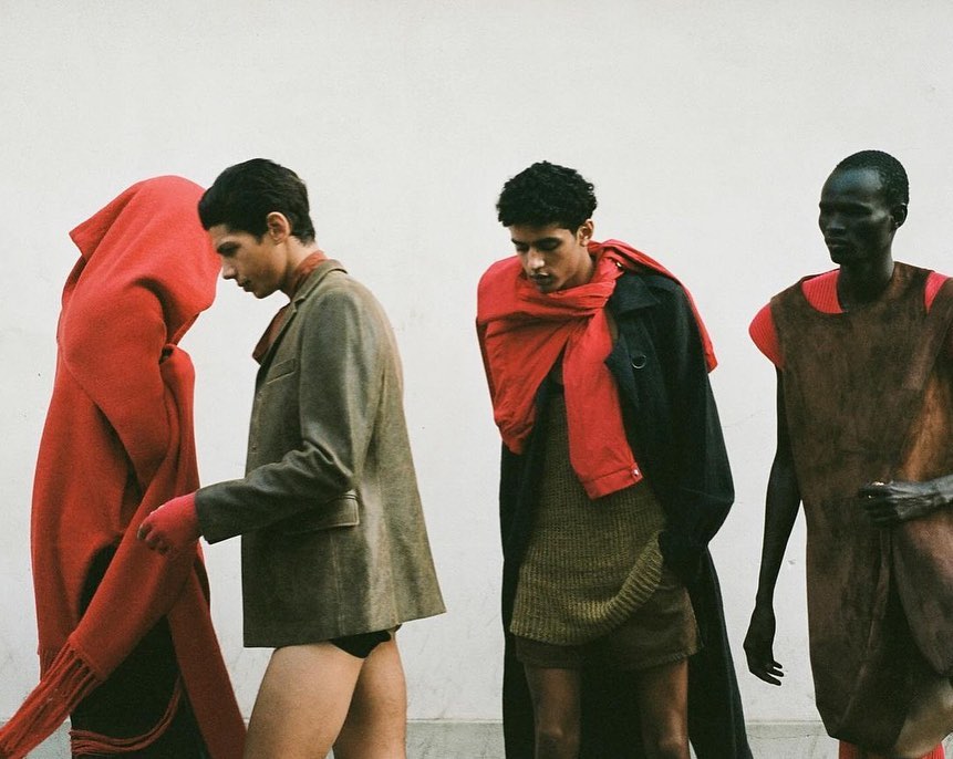 Fashion editorial shot of four men in a line wearing stylish outfits, set against a plain white background, showcasing modern clothing design.